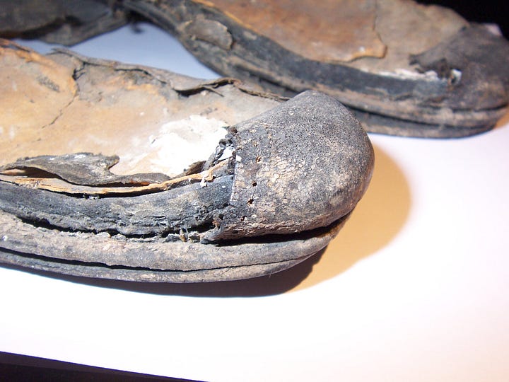 A pair of old shoes, partly decomposed, with patches of leather missing and flaked surface.