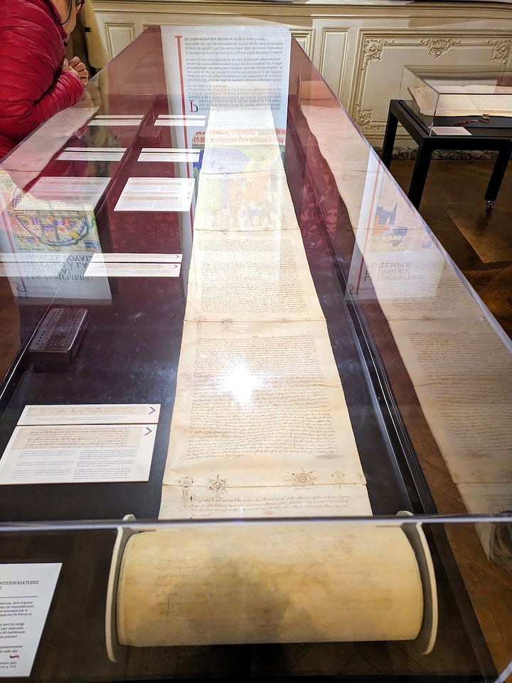Two views of museum vitrines, with handwritten documents on scrolls of parchment. The one to the right is significantly longer and the unrolled section is thicker.