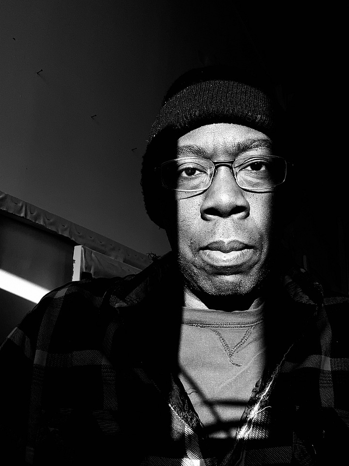 Two black and white photos of Tim McFarlane in his studio. Left: a shaft of light shows the artist's eyes and glasses and little else. Second photo shows the artist's full face, facing forward in a strong cylinder of light. 