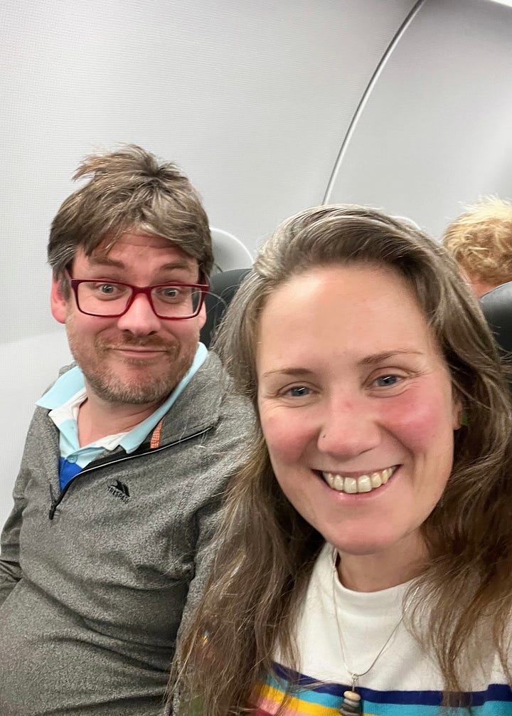 Image 1: H&E on the plane smiling at the camera. Both white with brown/grey hair. Photo of a cup of tea & a tin of red wine on a flight table