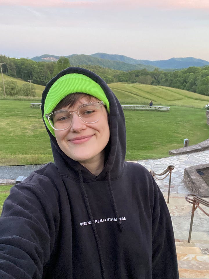 Shady standing in front of grassy hills and mountains in a hoodie and a dormatory house with blue sky and moon behind it. 