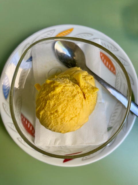 a ball of bright orange mango gelato in a clear bowl on a white plate with blue, red, orange, yellow and white leaves and a silver spoon and then the empty bowl afterwards