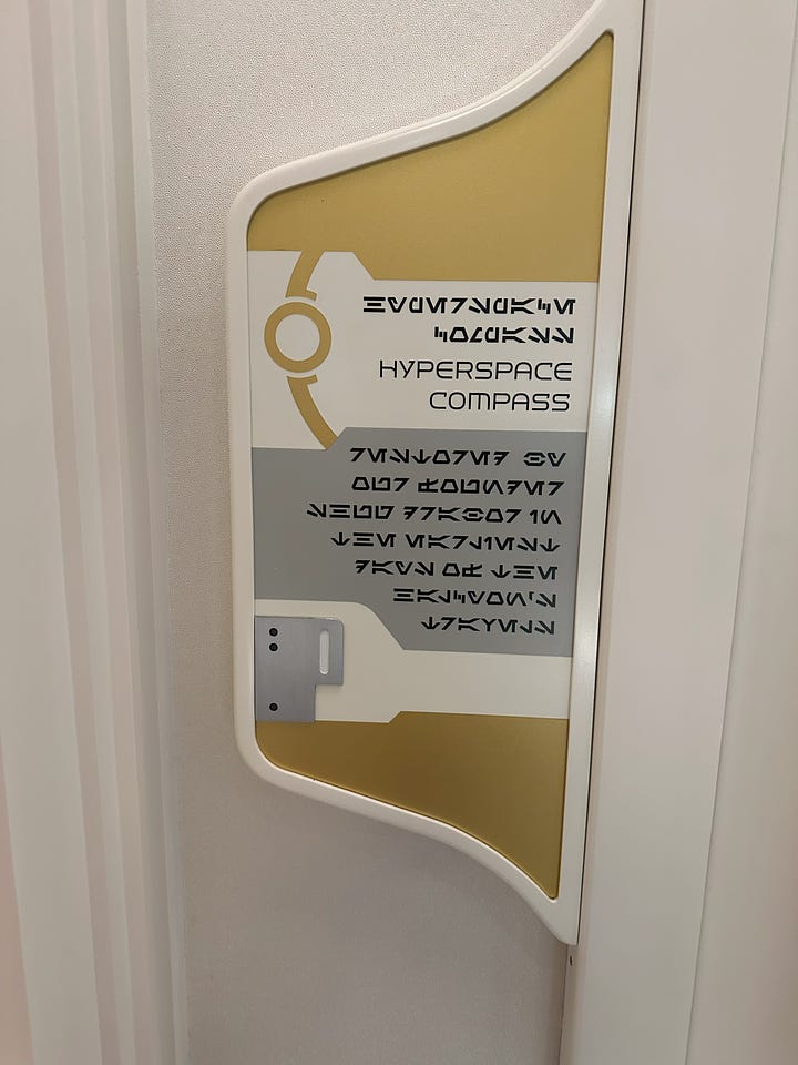 1st image: Hyperspace compass set into the wall, behind glass; 2nd image: A plaque. Header reads "Hyperspace Compass" in both English and Aurebesh. Below, in Aurebesh only: "Restored by our founder, Shug Drabor, in the earliest days of the Halcyon's travels"