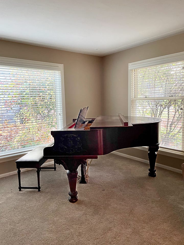 A baby grand piano stands in the corner of a room flooded with natural light. A toddler stands on the ground reaching up to touch the keys.