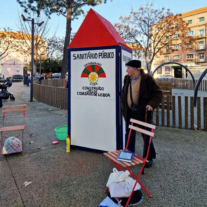 Foto 1: Un signore anziano, con indosso un cappotto scuro e un berretto, si trova accanto a un gabinetto pubblico bianco con un tetto rosso appuntito. Sul lato del gabinetto è presente una scritta in portoghese che recita "SANITÁRIO PÚBLICO INAUGURADO 2024 CONSTRUÍDO PELOS CIDADÃOS DE LISBOA", accompagnata da un disegno colorato che assomiglia a un sole sorridente. A sinistra del gabinetto ci sono due sedie di legno rosse e una bacinella verde. Lo sfondo mostra un ambiente urbano con alberi e edifici. Foto 2: Un cartello bianco con una sommità rossa e una cornice blu mostra la scritta "CASA DE BANHO DO POVO" in lettere nere. Sotto la scritta, c'è una piastrella bianca e blu che raffigura un gabinetto, con la scritta "WC" in piccolo in basso. Il cartello si trova all'aperto, con alberi spogli e persone sfocate sullo sfondo.