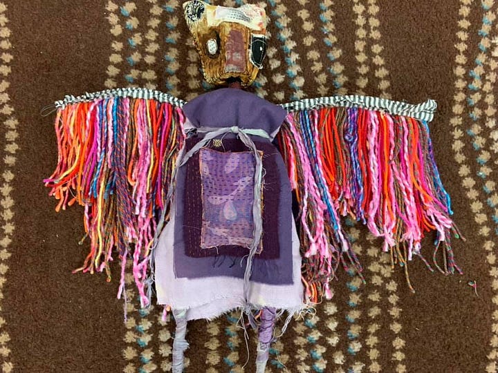 'Clockwise from top left, a plush doll with a brown face, a purple body and multicolored (primarily purple, pink and orange) feathers; the "not perfect" doll features a rabbit-eared figure with "not perfect" written over their mouth, they're sporting a yellow jumper with a big crimson heart on it; bottom, right, is a visual painting, "life," featuring two figures in Deveney's evocative "drip-like" style; and last, a doll with a bowling-pin-like body, with big sympathetic dark eyes, two big low ears that begin where earlobes would and a multicolored jumper on the body with knitted elements and a big red heart.