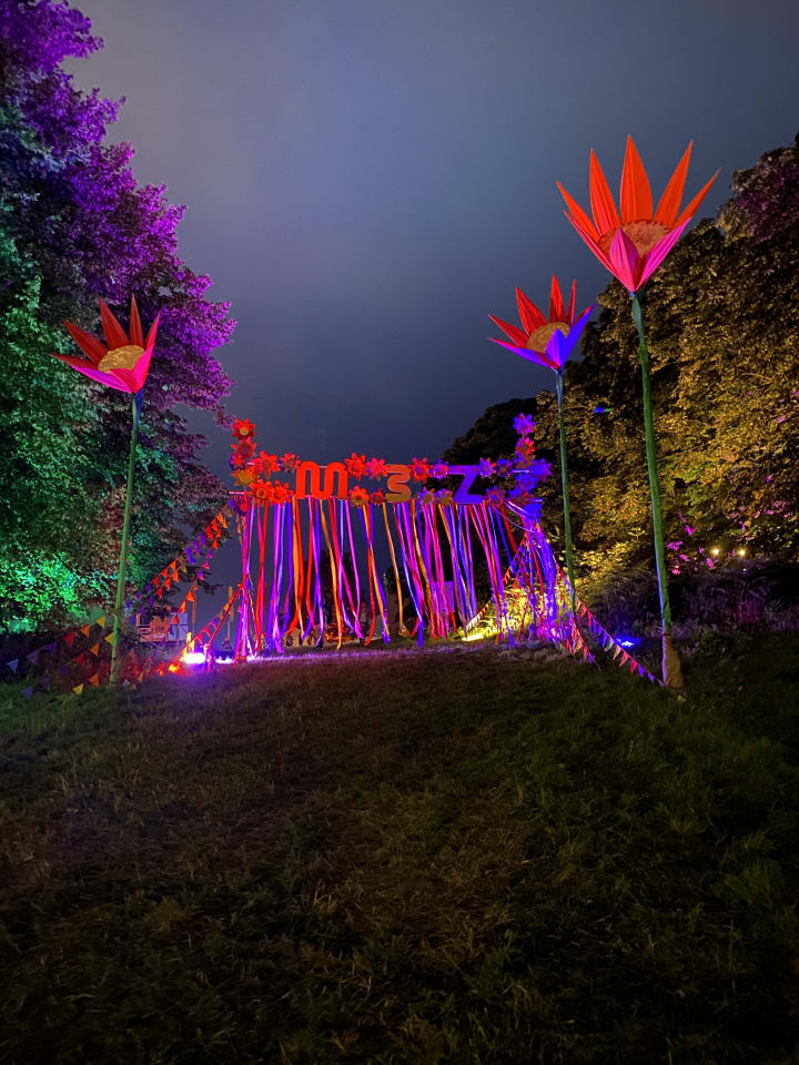 Artistic lighting at latitude festival (large latitude letters in a reflective metal lit up, multi-coloured strips leading into the zen area lit up with giant brightly coloured flowers leading to the entrance, lights strung up between trees in the wood and finally a kaleidoscope dome in gold