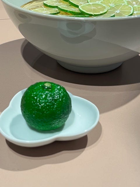 Image on the right is of a a plate of slices of a green, Japanese citrus fruit called Sudachi. On the left is a whole Sudachi, about the size of a lime, with a knobbly skin