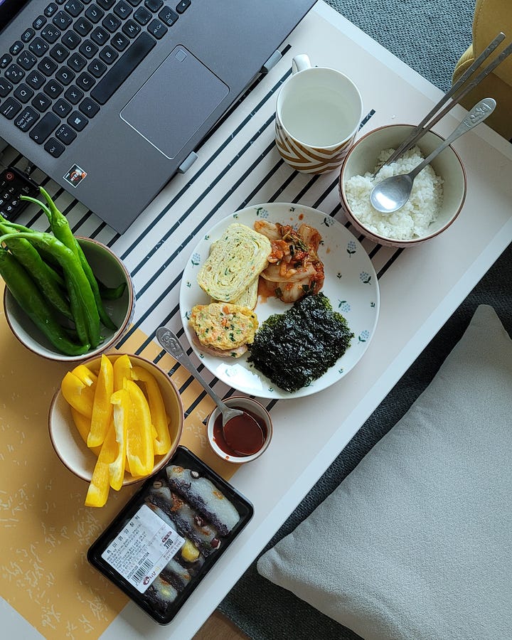 left: a photo of ji-youn's hand holding up a peeled mandarin orange, against the backdrop of the beach on jeju island. right: a birds eye view of dinner for one, with banchan, rice, and a laptop keyboard.