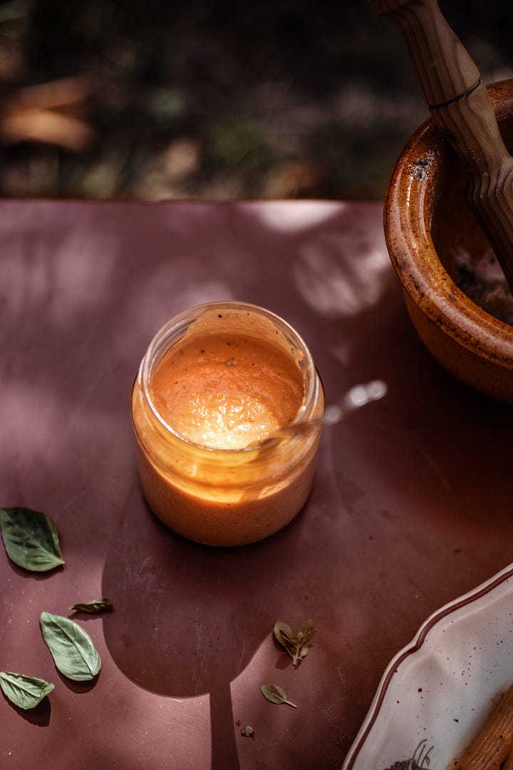 Creamy red pesto in a small glass jar on a red tablecloth, penne pasta with red pesto in an old fashioned pasta bowl with flowered edges on an outdoor table dappled with shadow from the trees and sunlight