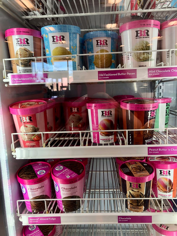 A brightly lit glass case with several different flavors of ice cream in pink and white containers, and a scoop of Turkey Day Fixin's ice cream in a white cup, with the BR/31 logo on front.