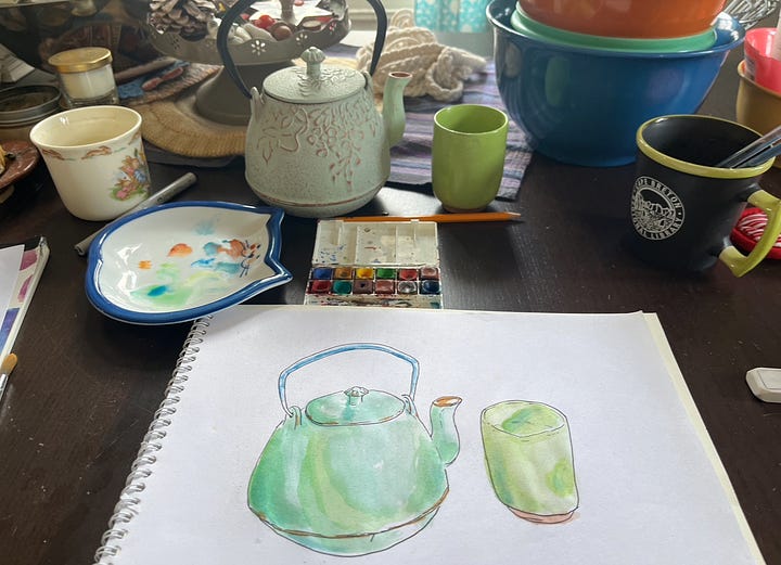 A pad of paper on my table with a black and white line drawing of a teapot and cup. The second image, the drawings are painted with watercolour