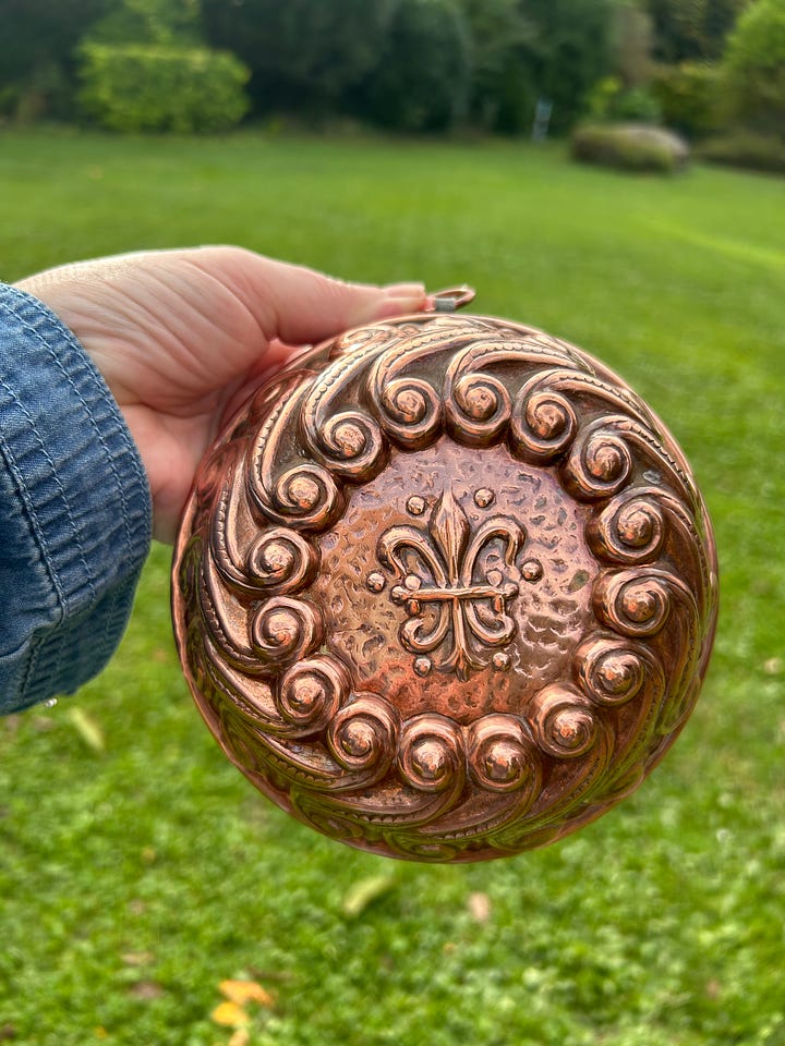 Karen holds a copper cake pan with an ornate swirl design and a fleur de lys accent. The results of the cake are displayed on a pretty table.