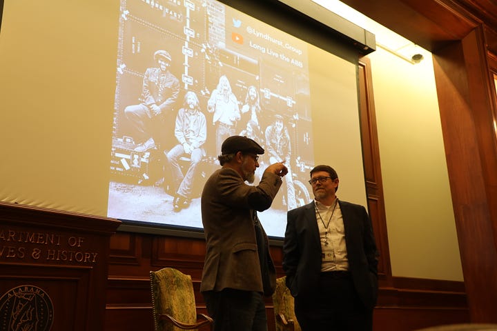 Bob Beatty (left) and Steve Murray (right) sit in chairs. Bob is wearing a hat, a tweed coat with elbow patches, a black Muscle Shoals Sound tshirt, blue jeans, and boots. Steve is in a gray suit with a white shirt. 