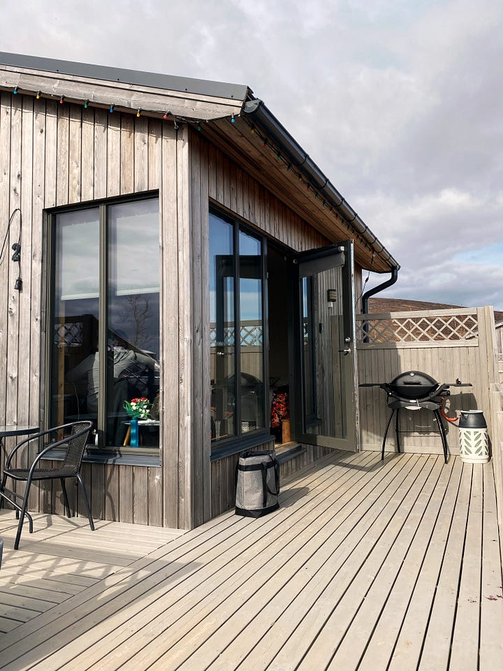 Our wood sided tiny house in the country with large windows seen from outside with a table, chairs and a barbeque.  Second image is the peaceful view across the empty fields in front of us under a blue sky. 