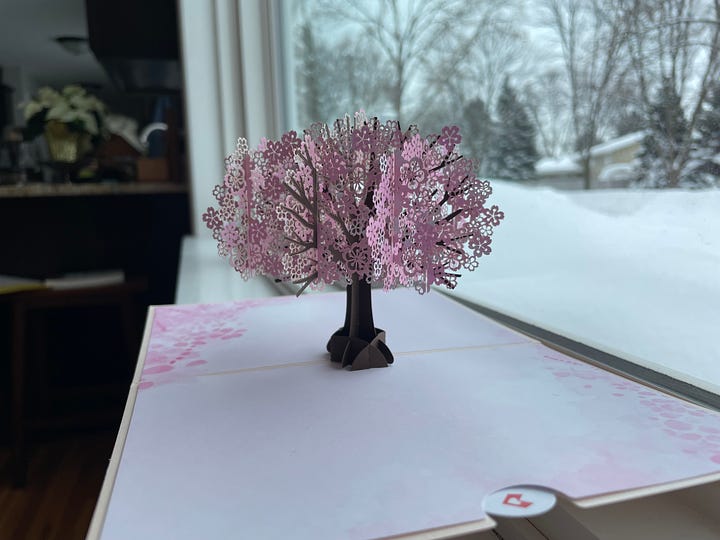 A pop-up cherry tree card featuring a quote by Madeleine L'Engle: "We turn to stories and pictures and music because they show us who and what and why we are."