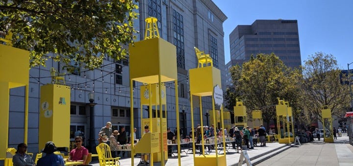 The first photo shows the gray barren plaza in front of the San Francisco Public Library. The second photo shows the same plaza filled with bright yellow columns containing exhibit interactives.