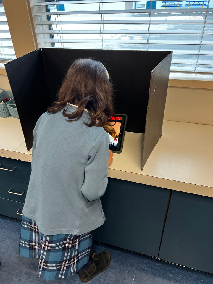 Photo of grade 1 student recording her video in a small cardboard divider. Her back faces the camera.