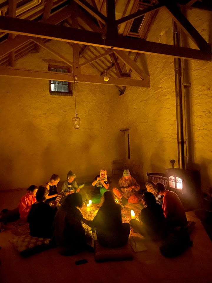 Sunset sky with a garden in the foreground, a blue shed to the right. A Black woman's hands holding a handmade cardboard loom with yellow wool. A floral mandala arrangement. A group of women sit in a circle in a barn. One woman is standing and gesturing widely (white, shoulder length brown hair, wearing a pink jumpsuit). A group of women sit in a circle lit only by firelight and candlelight