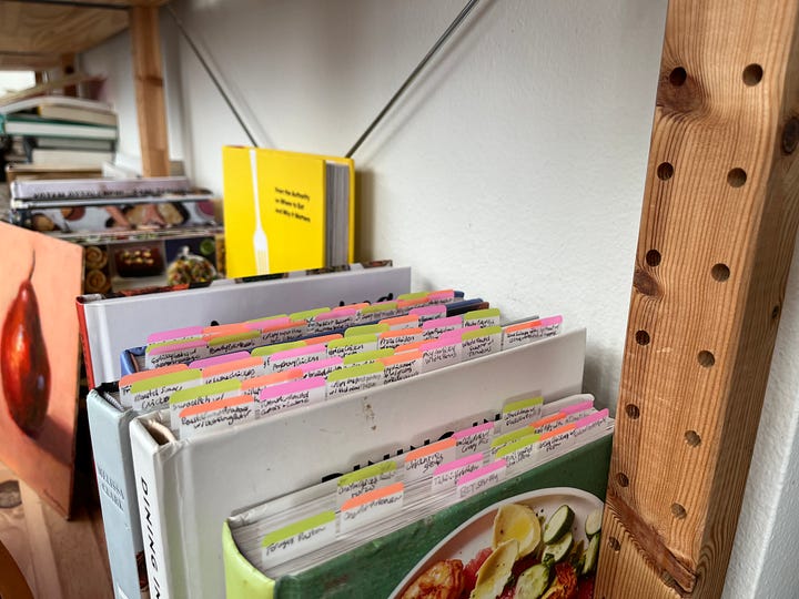 A photo of cookbooks on a shelf with Post-It tabs sticking up from the pages; a view of an onigiri mold with rice and salmon inside; a table with a pot of kabsa, a bowl of greens, and a small bowl of yogurt; a view of a pan with cooked harissa chickpeas and pieces of feta.