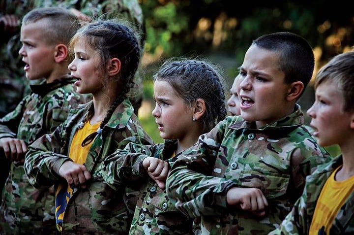 Pictures show young children being taught how to use rifles by Azov members with Swastika tattoos.
