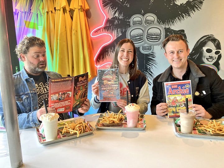 Jordan Morris, Amy Chase, and Ryan Cady sitting at a diner with burgers, fries, and milkshakes. Each is holding a copy of Pop's Chock'lit Shoppe of Horrors from Archie Comics. Amy in her Halloween costume, with a full-face zombie mask inspired by the fungal Clickers of The Last of Us.