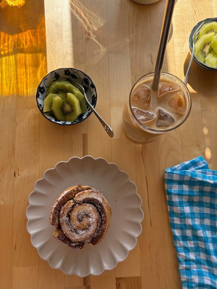 Four sugar-topped cinnamon scrolls on a wire rack. A cinnamon scroll on a plate with sliced kiwi fruit and an iced latte