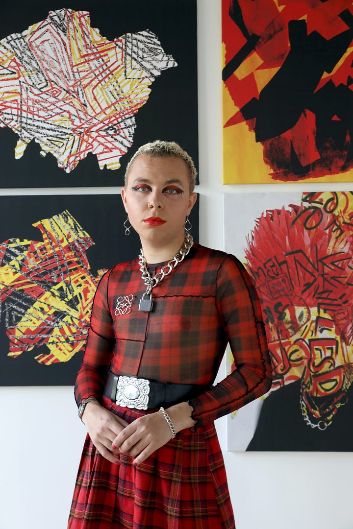 Warren wears a red and black tartan sheer shirt. He had strong red and black eyemakeup, red lipstick and short blonde curly hair. He wears a thick silver padlock chain necklace, a black belt with a silver buckle and stands in front of his artwork