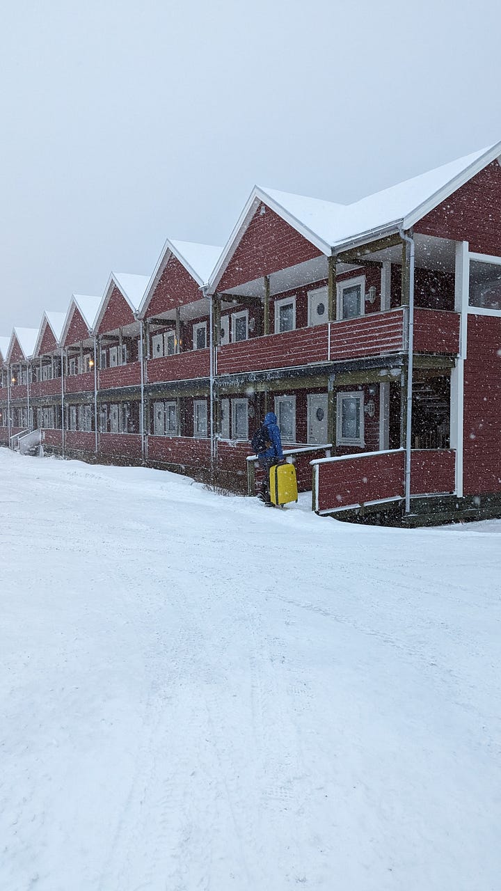 A line of cherry-red wooden cabins sits in the falling snow, and a woman stands at the end of a snow-covered pier, the fjord and snow-topped mountains behind her upstretched arms.
