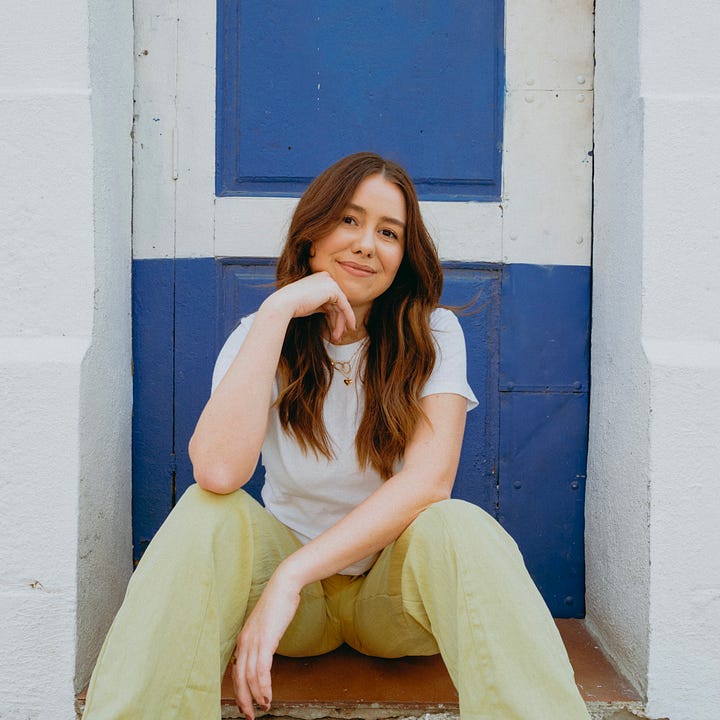 Headshots of Freelance Copywriter, Sawyer Wilson. In the image on the left, she is smiling directly at the camera, and in the image on the right, she is seated in green pants in front of a blue door, with her chin resting on her hand, smiling at the camera.
