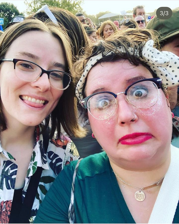 Holly, non binary person smiles while wearing a striped shirt glasses, butterfly clips and glasses with Janet, also smiling, wearing a black shirt and glasses. Janet stands wearing a blue and yellow patterned shirt and black pants stands next to Holly wearing a white patterned dress, a green wig, and antler flwer crown.