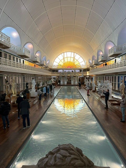 Wide shot of swimming pool gallery; Hommes Dames pool entrance; white marble sculpture of bather; plaster of Paris sculpture of torso with red mushroom cloud in place of the head