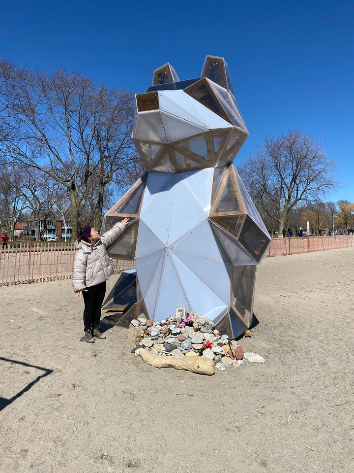 Girl with giant raccoon statue. House shaped wooden building with neighbourhood names on the side.