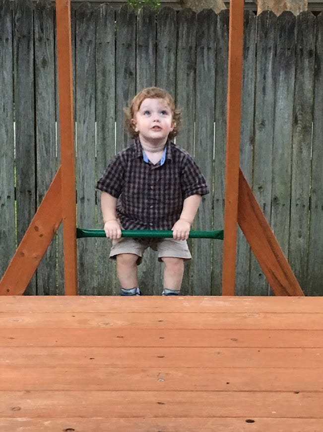 The image shows a young child standing on a wooden deck, gripping the rungs of a ladder that leads up to a playset. The child is looking upwards, perhaps at someone or something not visible in the photo. He is wearing a plaid shirt, shorts, and sandals, with a backdrop of a wooden privacy fence. The moment captured is tranquil, likely a snapshot of playtime or exploration. Second picture:  The image features a young boy sitting at the top of a green plastic slide, his hands gripping the sides as he prepares to slide down. He wears a plaid shirt and shorts, and his expression is one of concentration and cautious excitement. In the background, another child is visible, standing on a wooden deck. The setting suggests a playful and curious atmosphere in a residential backyard, with a wooden fence surrounding the area.