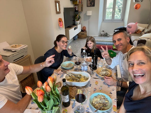 people of all agres sitting around a table in italy, drinking wine, smiling, talking, eating pork shank and potatos