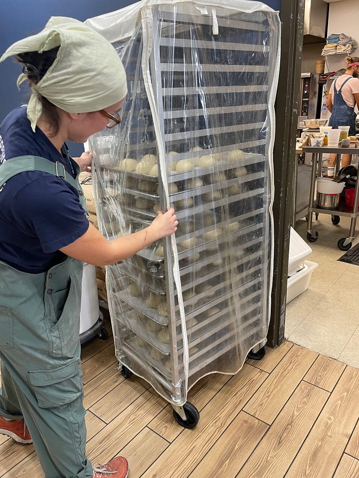 Beverage cans are lined up for sale. A worker wearing overalls and bandana over her hair tends to a cart of baked products.
