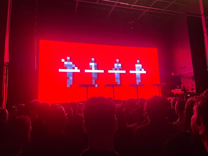 First photo of Lyss against a backdrop of red light, wearing black lipstick, a black choker, and a black tank top. Second photo of Kraftwerk live in concert, against a red background. Third photo of a donut with rainbow sprinkles. Fourth photo of a pitbull with a donut toy in her mouth, laying happily on a bed.