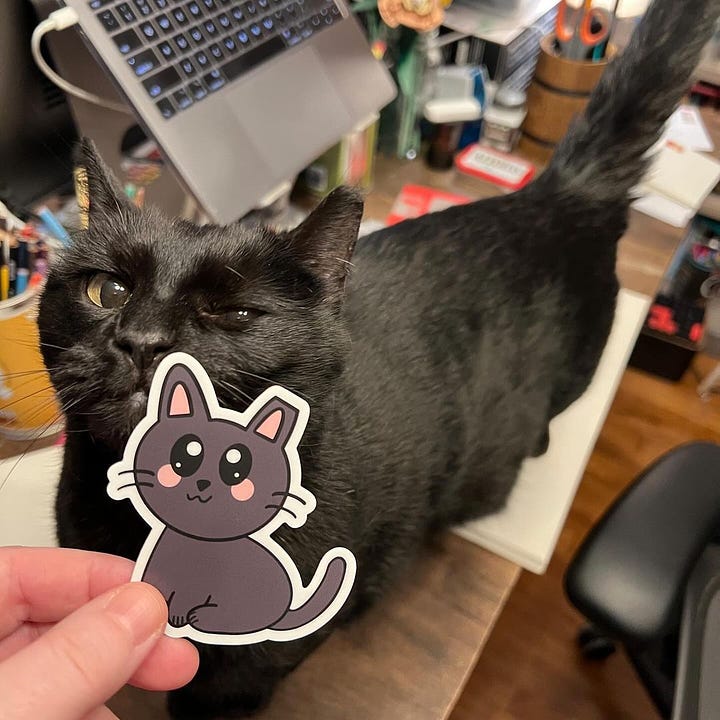 A grey and white tabby cat lounges on top of a sketch book and checks out the sticker of himself that his mom is holding. A black cat stands on top of a desk and checks out the sticker of himself that his mom is holding.