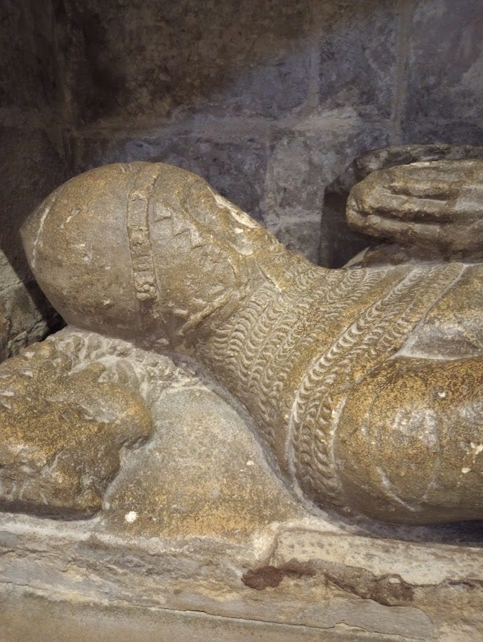 Tomb in wall, with canopy, featuring lifesize effigy of medieval knight with chainmail and helmet.