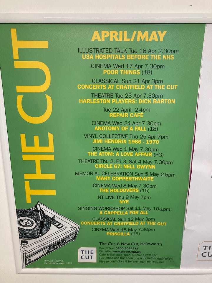 Photo of Vicki Lesley and Pete Wilkinson in front of a brick wall / green poster for events in May at The Cut in Halesworth