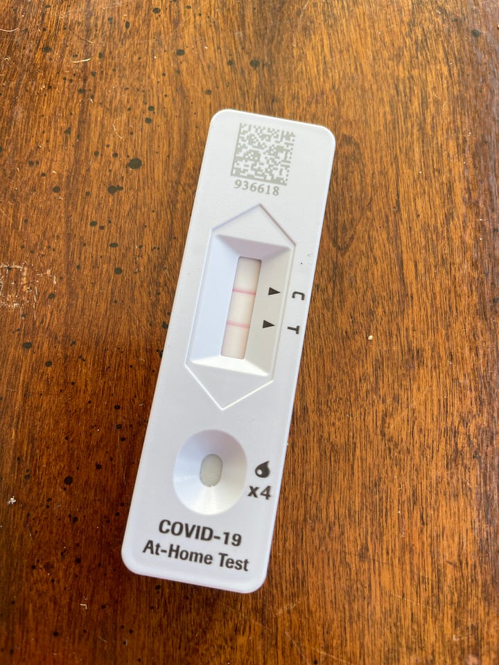 A positive covid test rests on a wooden table.  A delicious pot of chicken soup with the lid partly off.