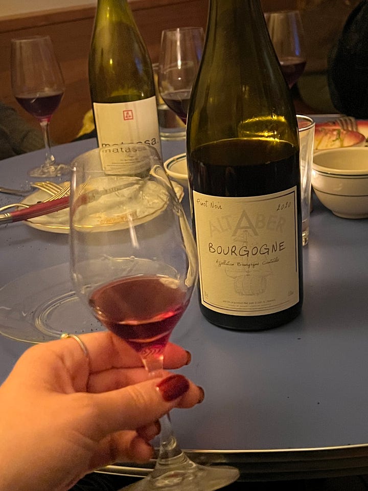 Glass of red wine on a blue table with a couple bottles of wine in the background. second picture: Two men holding sitting at a table, each holding a bottle of wine with wine glasses and food on the table in front of them.  