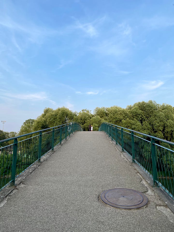 1. Top left.  image I took of a pedestrian bridge in France. Lush green trees are in the background, and there is a soft blue sky with hints of white clouds— in almost crescent-like formations. If you look closely, you can see two people walking together.. The railings are green, and the bridge is concrete. 2. Top Right. Image of a Keith Haring collage at Piedmont Park. Haring’s hair is purple, and he’s wearing red glasses and a yellow shirt. The vendor also sells t-shirts- mostly neutrals and Black Power Africa jewelry— behind the collage. A man is standing near the Keith portrait wearing white pants, a gray shirt, and a black watch. I’m walking with Jarvis, unseen, telling them about a dream Malcolm and I have.  3. Bottom Left. Image of a brown paper bag containing one of my favorite meals. Written on it says, “From We, With Love,” with a wide heart below it. Surrounding it are the names Eugeny, Olya, and Alex.  4.Bottom right.  Image of a collection of smiley faces while walking through the Denmark airport. A variety of faces drawn in white paint with a sharp dark blue background. One smile has a mirror that also has neon typography in it, it says “expect a miracle.” 