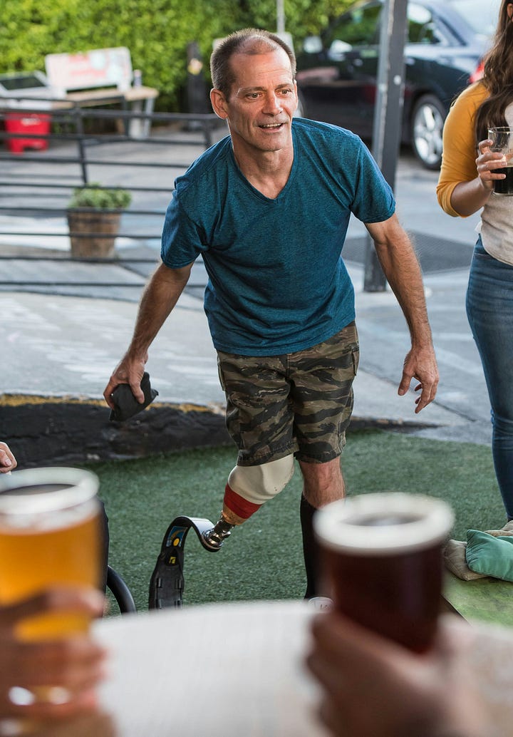 Left: Short-haired man with a prosthetic leg wears a blue t-shirt and camouflage shorts. Right:  A short-haired man using a wheel chair positions himself by an open car door. He wears a peach-colored shirt and tan pants.