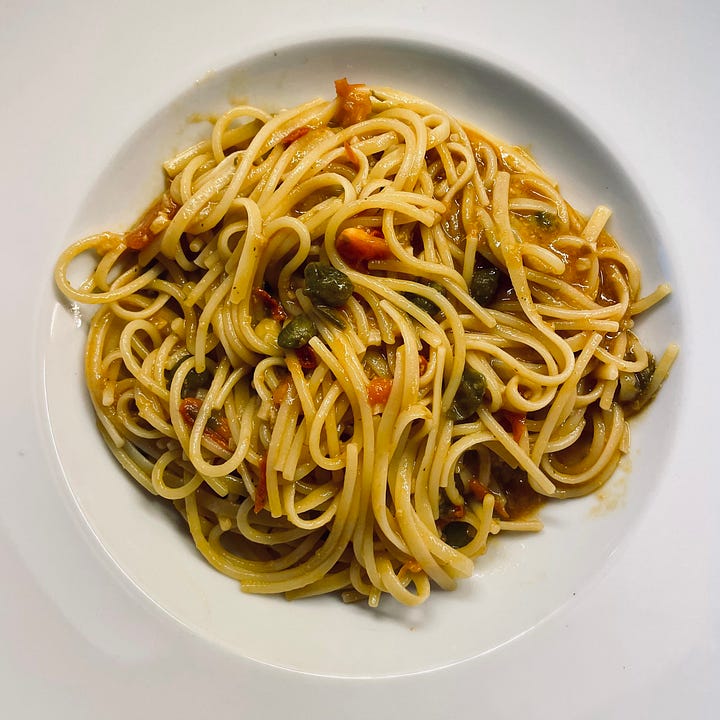 A plate of spaghetti with a rich tomato-based sauce, capers, anchovies, garlic, and chili, topped with fresh baby tomatoes and a drizzle of olive oil, served on an outdoor balcony.