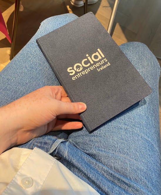 Red-headed woman smiling in front of a Social Entrepreneurs Ireland banner, and a notebook embossed with Social Entrepreneurs Ireland 