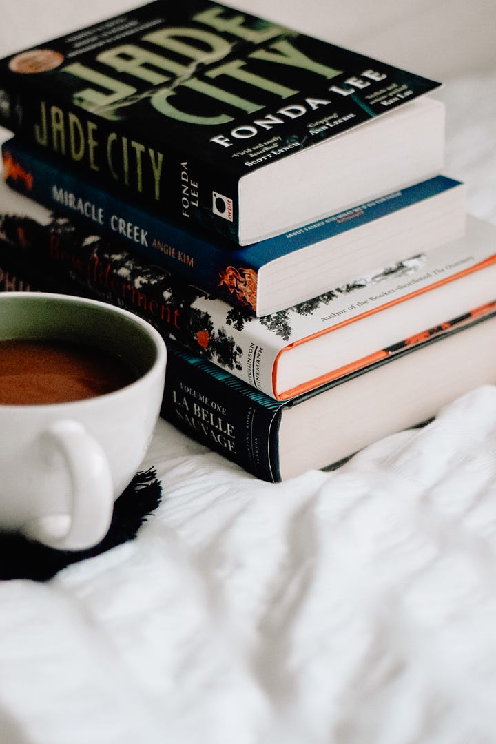 Stack of paperback books on a cosy white duvet, with a cup of coffee.