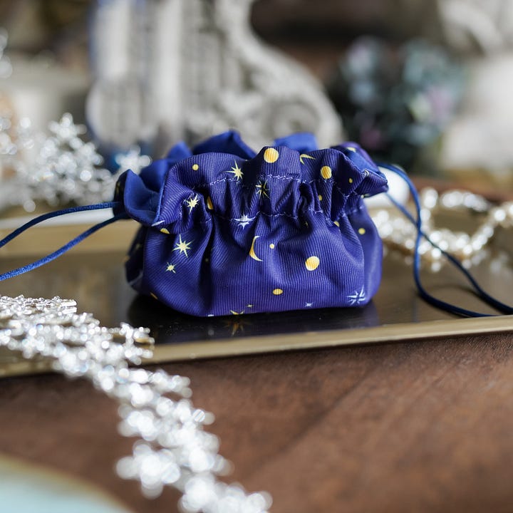 Two photos of the bag of infinite space drawstring bag. The bag is dark blue and scattered with moons, stars, and compasses. The second photo shows the bag's interior with a piece of golden jewelry in the pockets.