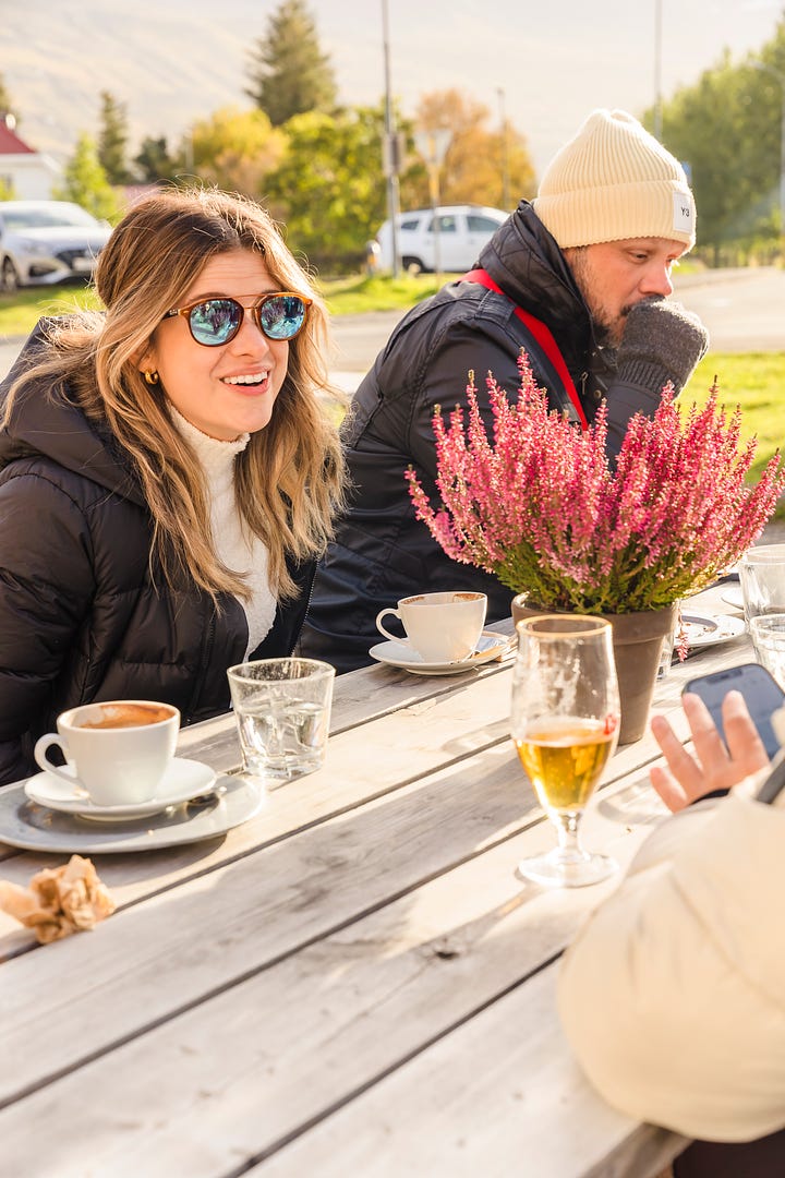 Photo of woman standing along a black sand beach to look at shells and pebbles; woman smiling sitting on a large boulder along a river filled with glacial melt and icebergs with a bridge in the distance; woman laughing and talking at an outdoor picnic table with a group of friends having coffee; selfie photo of two woman smiling wearing sunglasses and winter clothes.