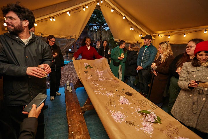 Two more photographs from the Glimpse Farewell. On the left is a zoomed out shot of the Glimpse timeline drawn on the brown paper table cloth. One of the Glimpse team members is holding the end of the cloth, as if about to roll it up. Guests are standing around her. The top of the tent is in shot, and it's decorated with large bulbed fairy lights. On the right is a photograph of Glimpse guests standing around the fire. James is about to throw a piece of the brown paper table cloth into the fire pit. Someone else is stocking the fire. It's dusk.